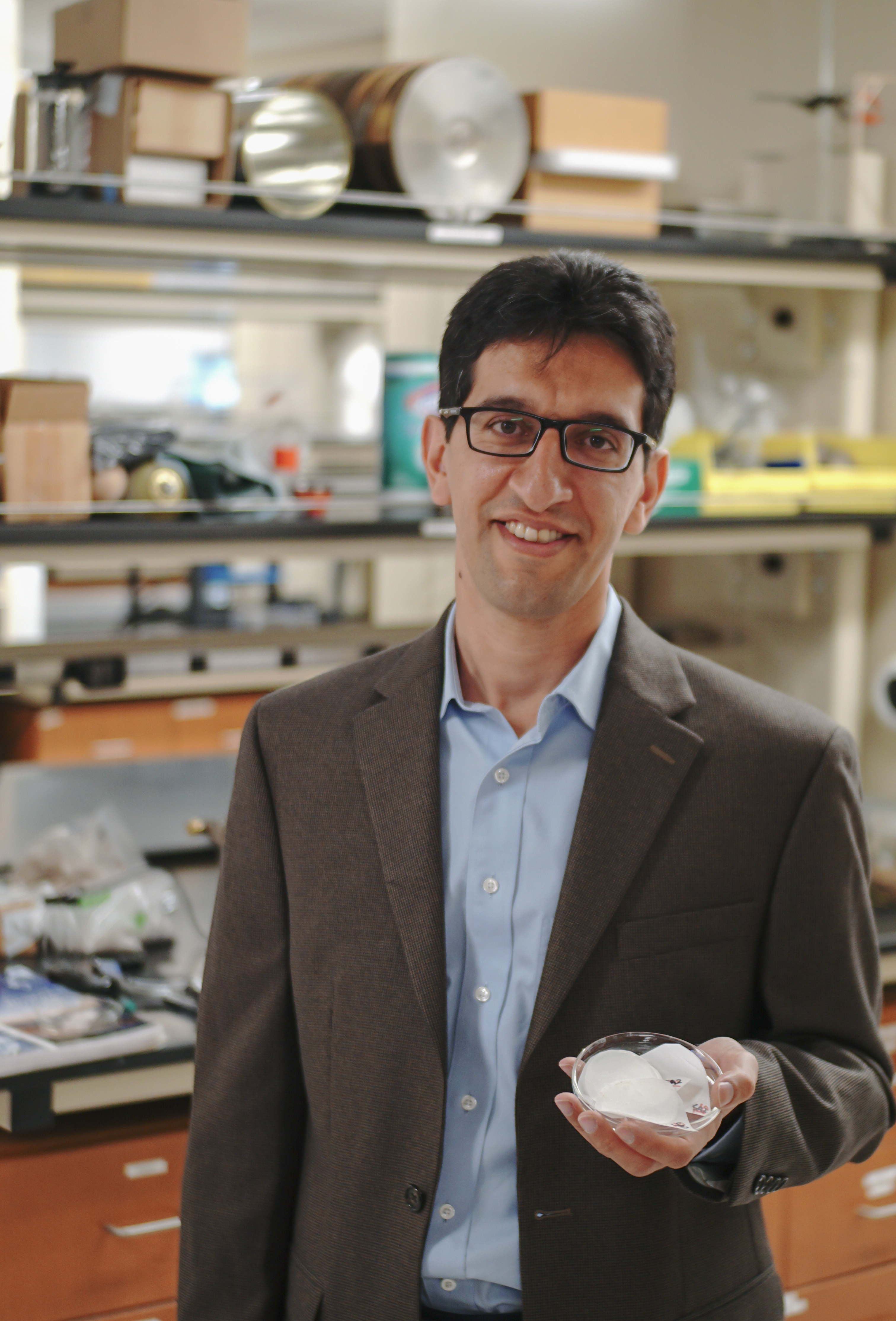 Man, standing in a lab, holds a Petri dish. 