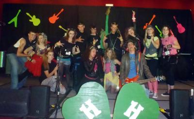 A group of 13 young people dressed as rockers pose in various positions that include extended tongues and extended fingers making the "peace" sign.