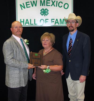 Two men and a lady, in a brown dress, standing in front of the New Mexico 4-H Hall of Fame sign.