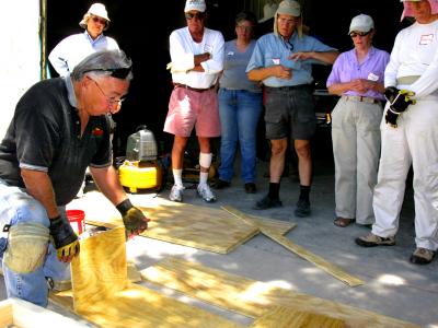 Six people watch in the background as a kneeling man fits plywood components together in an outdoor work area.