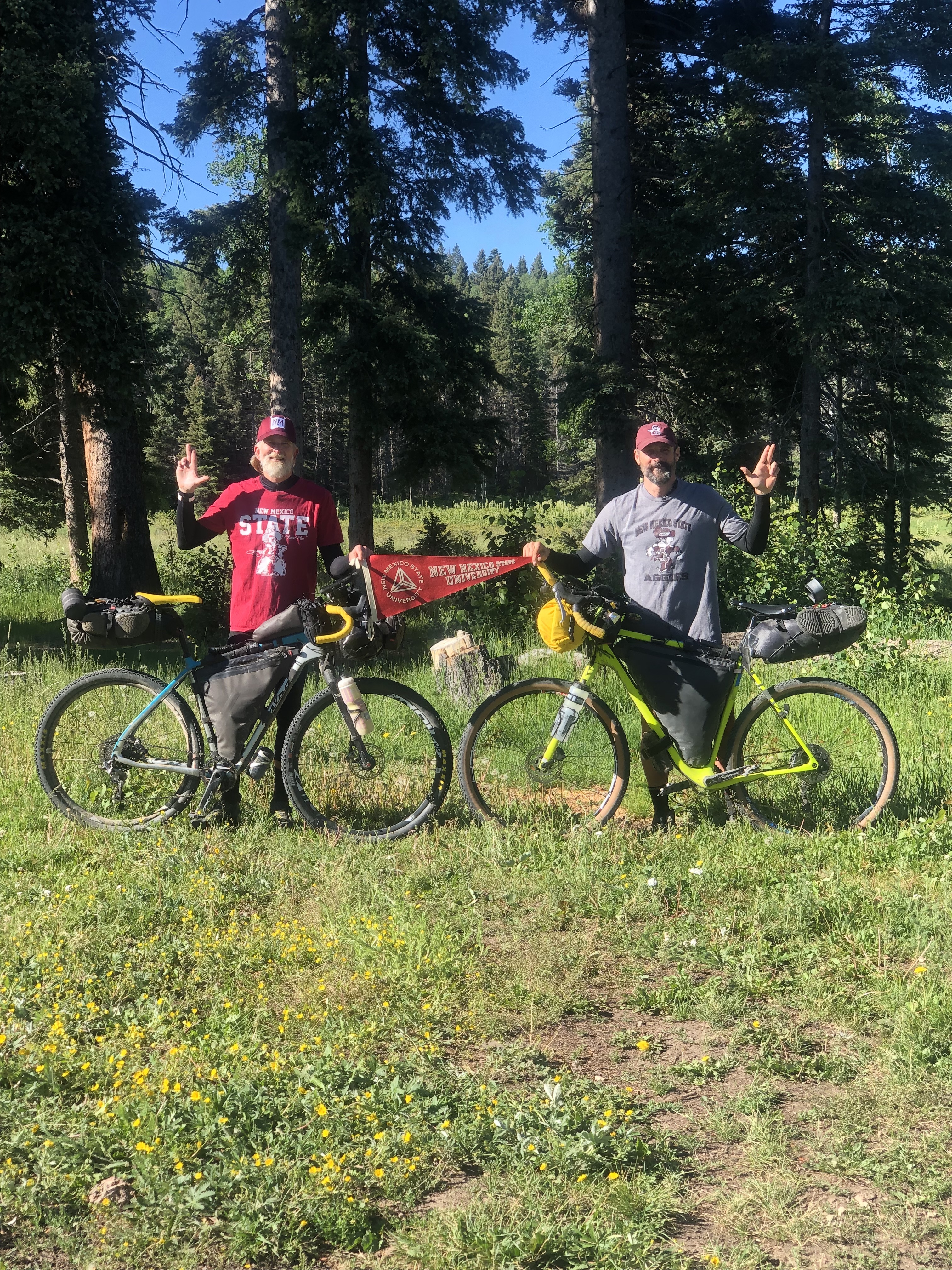 Two men standing with their bikes holding a NMSU pennant.
