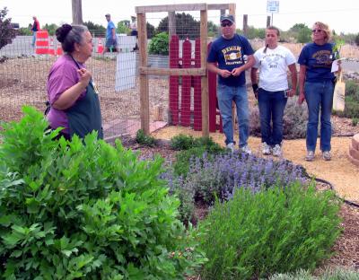 Woman with green apron standing in garden talking with three people.