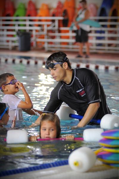 A swim instructor stands in the water surrounded by three elementary school children, one of whom is using a floating device.