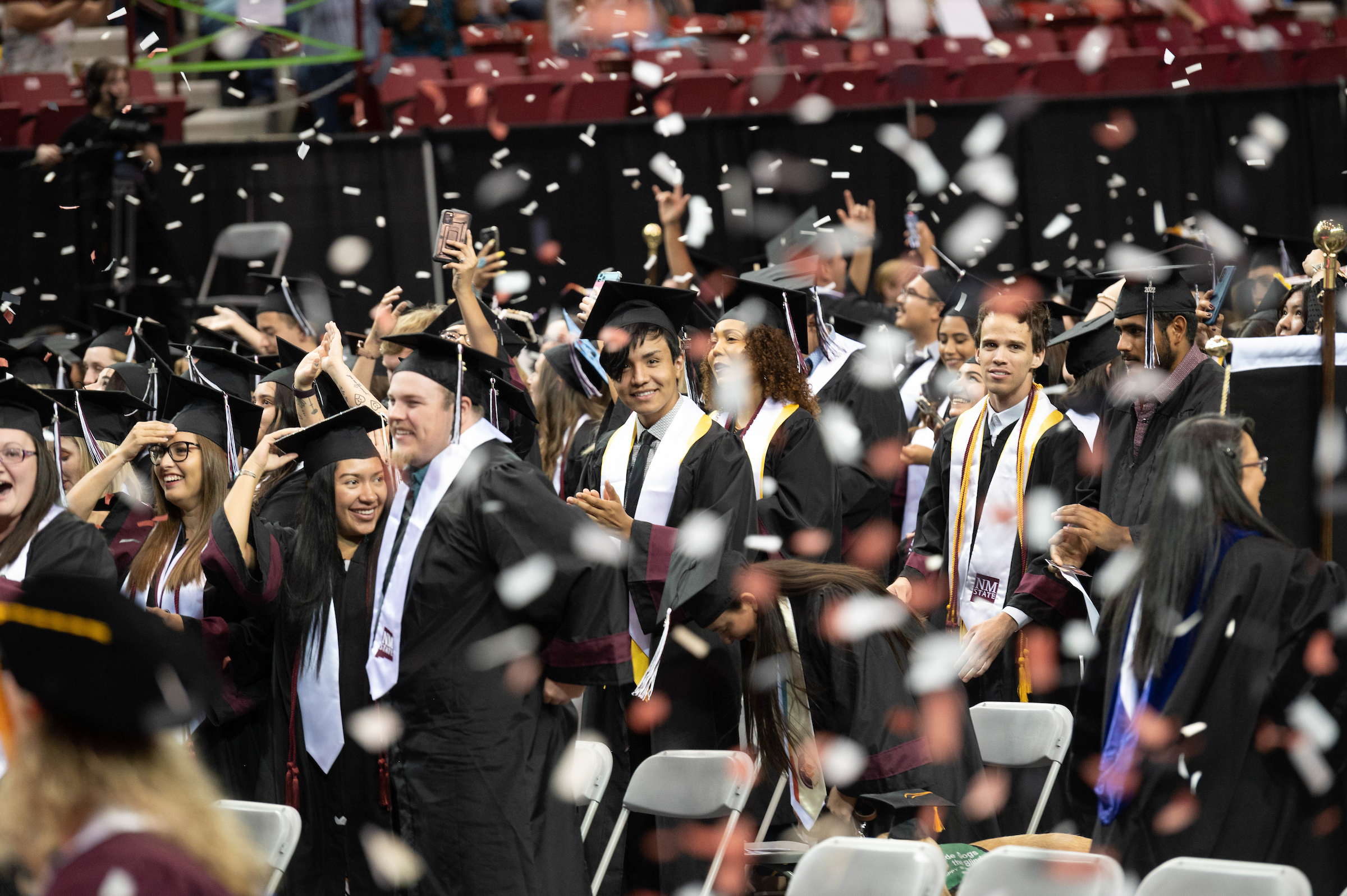 Men and women stand and celebrate as confetti flies at a commencement ceremony. 