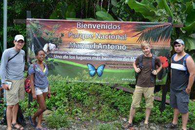 Four young people stand at the welcome sign for Manuel Antonio National Park in Costa Rica with dense jungle vegetation in the background.